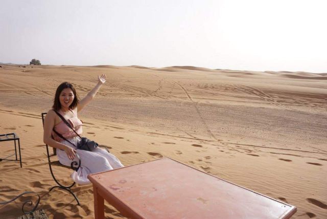 Smiling traveler waving from a chair on the golden sand dunes of Erg Chebbi, Merzouga, Sahara desert