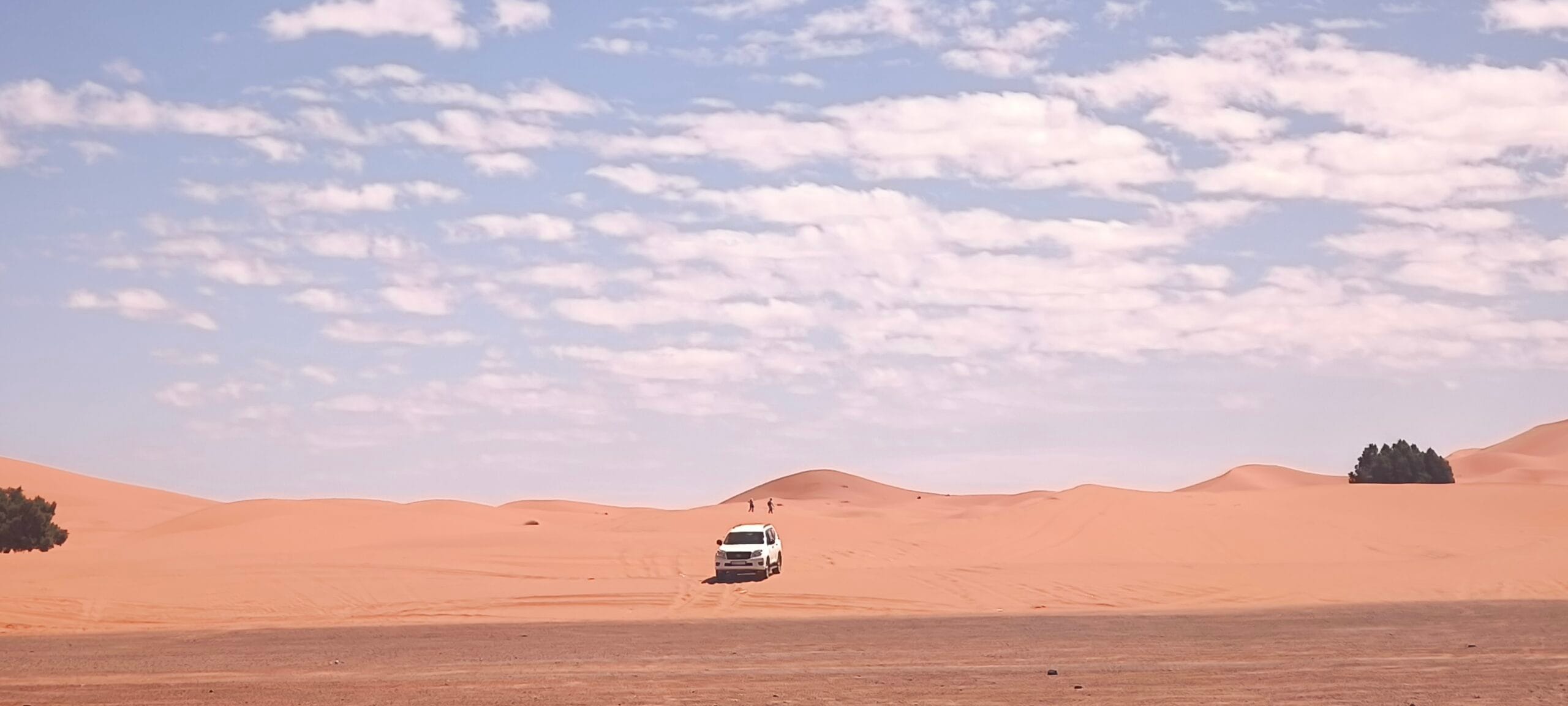 A 4x4 off-road vehicle navigating steep golden sand dunes in the Sahara Desert of Morocco, kicking up a trail of sand.