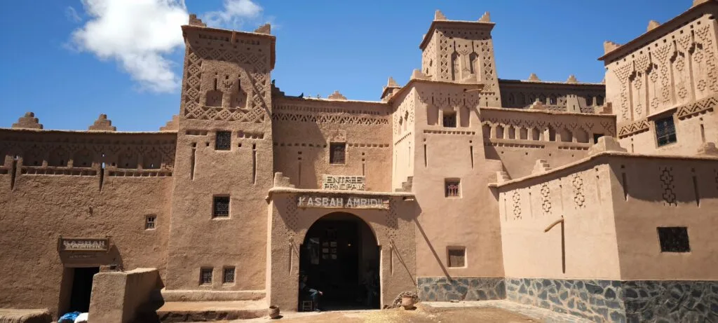 Majestic earthen towers of Kasbah Amridil rising above the lush palm oasis of Skoura in southern Morocco, with traditional pisé mud-brick architecture glowing in warm sunlight against a clear blue sky and the distant Atlas Mountains on the horizon.
