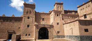 Majestic earthen towers of Kasbah Amridil rising above the lush palm oasis of Skoura in southern Morocco, with traditional pisé mud-brick architecture glowing in warm sunlight against a clear blue sky and the distant Atlas Mountains on the horizon.