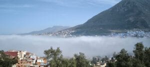 Aerial view of the blue medina of Chefchaouen in northern Morocco, partially veiled in soft morning fog and low clouds, with the iconic cobalt-washed rooftops and minarets emerging through the mist against the backdrop of the Rif Mountains.