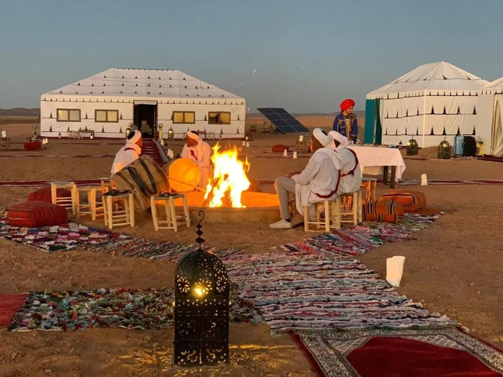 Travelers relaxing around a campfire at a Sahara desert camp in Morocco, with traditional tents and lanterns creating a warm evening atmosphere.