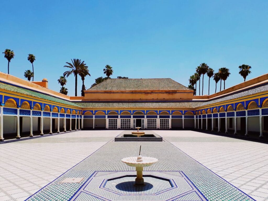 The ornate central courtyard of Bahia Palace in Marrakech, Morocco, featuring intricate zellij tilework, carved cedar wood ceilings, and lush orange trees surrounding a central fountain, bathed in warm afternoon light.