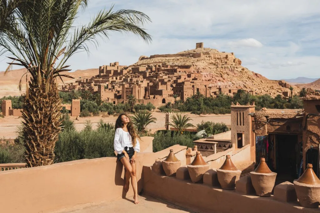 A happy young woman smiling while visiting Aït Benhaddou, the ancient UNESCO World Heritage ksar (fortified village) in Morocco, with its iconic earthen clay architecture and desert landscape in the background.