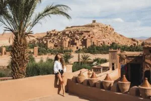 A happy young woman smiling while visiting Aït Benhaddou, the ancient UNESCO World Heritage ksar (fortified village) in Morocco, with its iconic earthen clay architecture and desert landscape in the background.