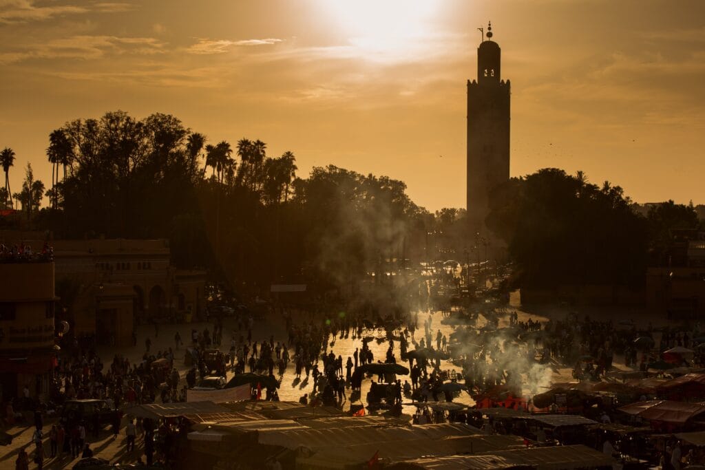 Vibrant Djemaa el-Fna square in Marrakech, Morocco, bathed in warm golden sunset light, with smoke rising from open-air food stalls, crowds of locals and travellers filling the plaza, and the iconic Koutoubia Mosque minaret rising above the rooftops in the background.