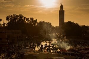 Vibrant Djemaa el-Fna square in Marrakech, Morocco, bathed in warm golden sunset light, with smoke rising from open-air food stalls, crowds of locals and travellers filling the plaza, and the iconic Koutoubia Mosque minaret rising above the rooftops in the background.