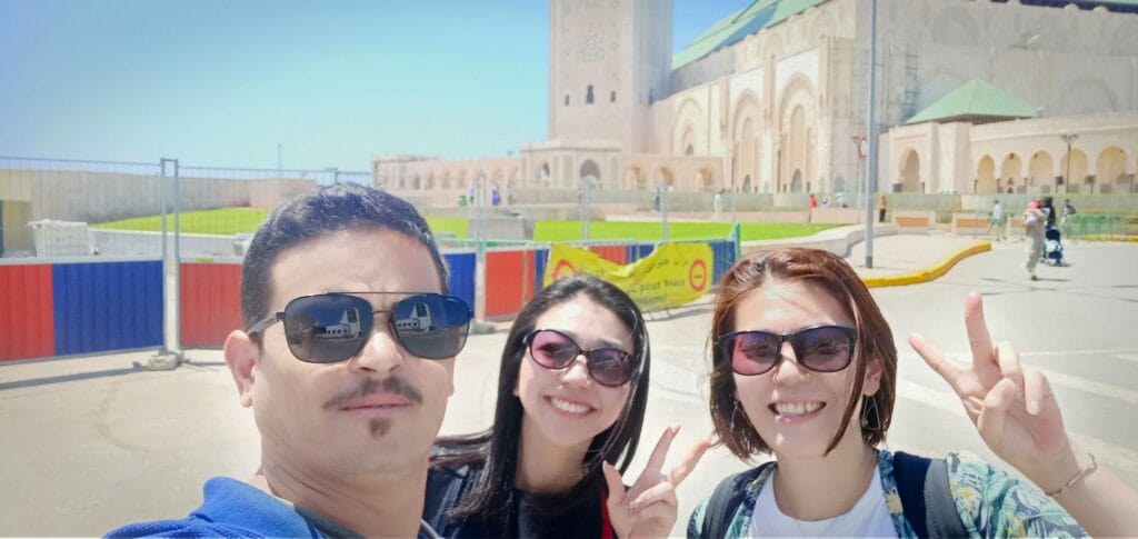 A Moroccan tour guide named Sabir with a group of Japanese tourists outside the grand Hassan II Mosque in Casablanca, Morocco, with the mosque's iconic minaret and ornate architecture visible in the background.