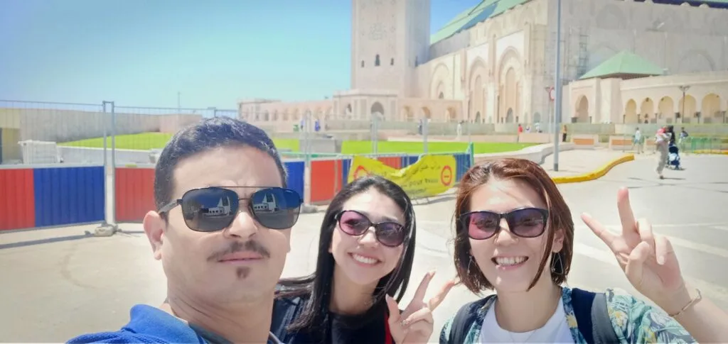 A Moroccan tour guide named Sabir with a group of Japanese tourists outside the grand Hassan II Mosque in Casablanca, Morocco, with the mosque's iconic minaret and ornate architecture visible in the background.