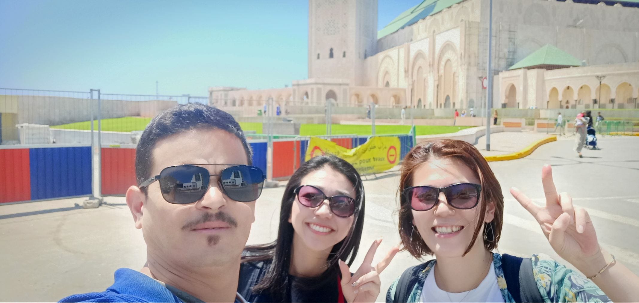 A Moroccan tour guide named Sabir with a group of Japanese tourists outside the grand Hassan II Mosque in Casablanca, Morocco, with the mosque's iconic minaret and ornate architecture visible in the background.
