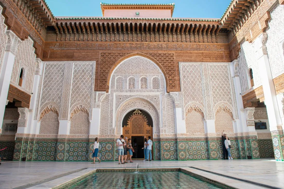 Interior courtyard of Ben Youssef Madrasa in Marrakech, Morocco, featuring intricate zellige tilework at the base, elaborately carved stucco walls with geometric and floral patterns, dark cedarwood screens and archways, and a central marble fountain reflecting the ornate façade under a clear blue sky.