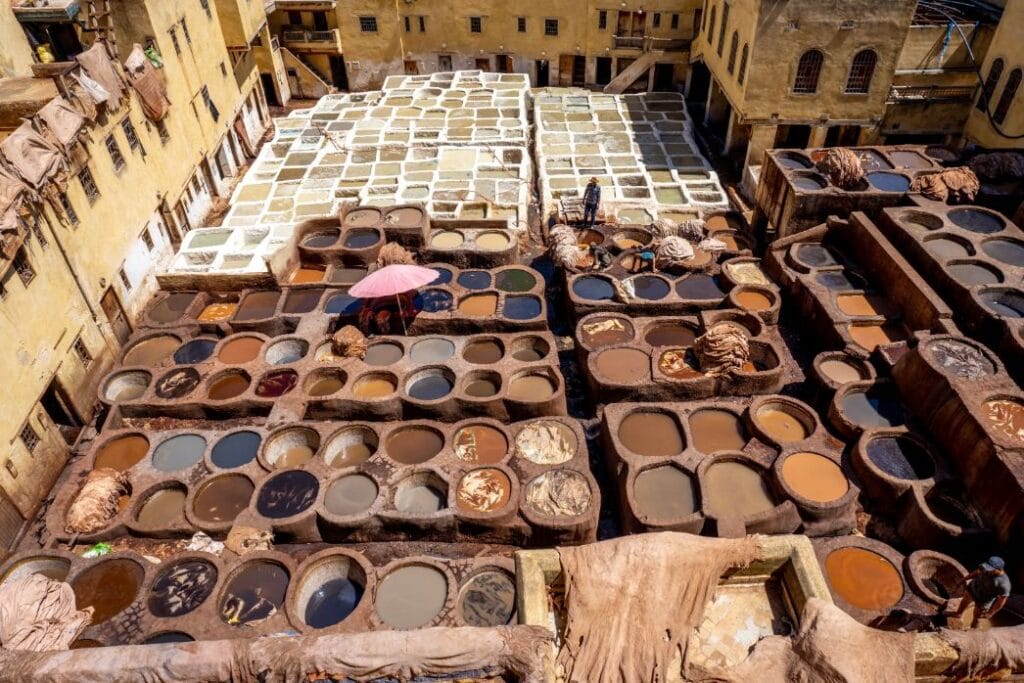 An aerial view of the iconic Chouara tannery in the medina of Fez, Morocco, revealing a mosaic of round stone vats filled with vibrant natural dyes in shades of saffron, poppy red, and indigo, surrounded by workers hand-dyeing hides using centuries-old techniques.