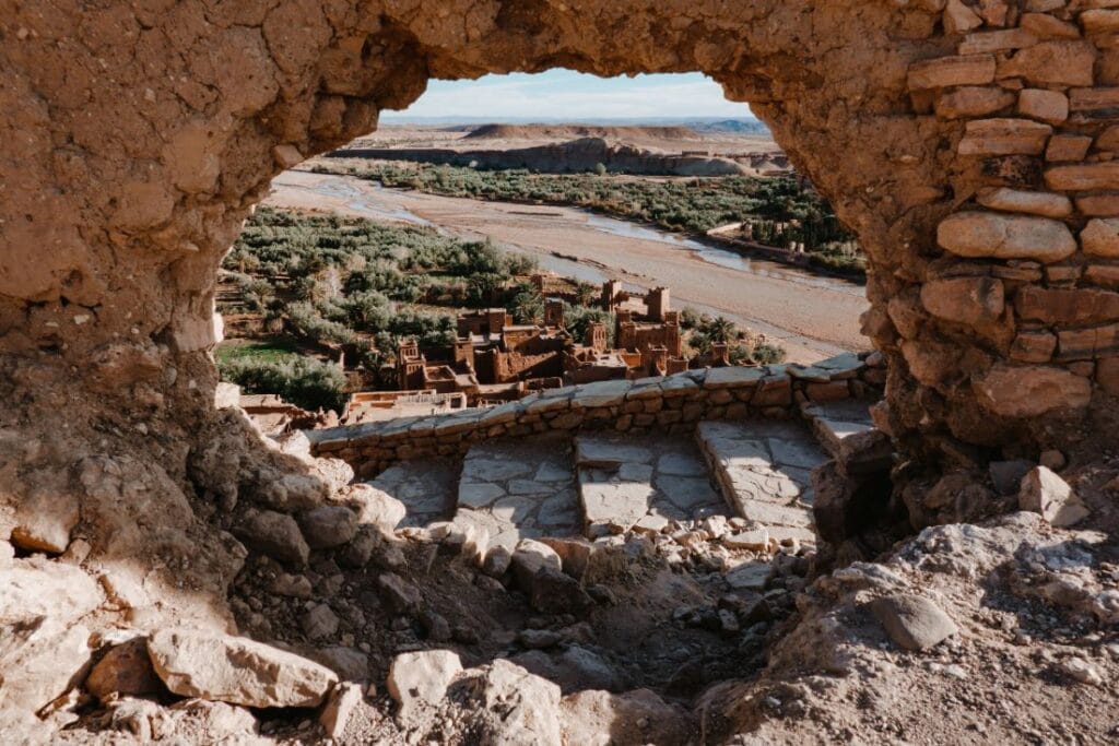 A panoramic view of the ancient fortified ksar of Aït Benhaddou, a UNESCO World Heritage Site in southern Morocco, with its iconic earthen clay kasbahs and towers rising above the Ounila River valley against a clear desert sky.