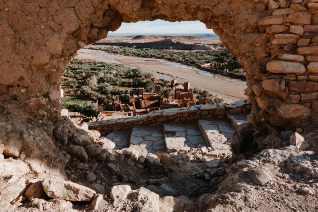 A panoramic view of the ancient fortified ksar of Aït Benhaddou, a UNESCO World Heritage Site in southern Morocco, with its iconic earthen clay kasbahs and towers rising above the Ounila River valley against a clear desert sky.